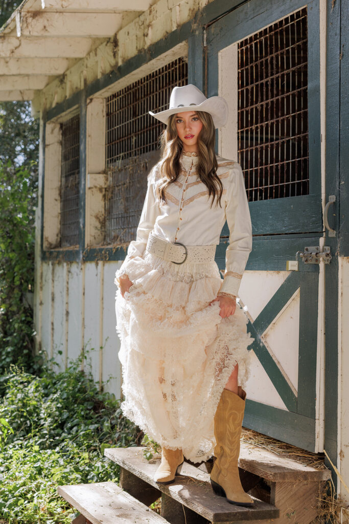 Senior girl wearing a white cowboy hat, vintage lace western blouse, tiered lace skirt, and cowboy boots, posing in front of a rustic barn door during a country western style portrait session in Carmel California 