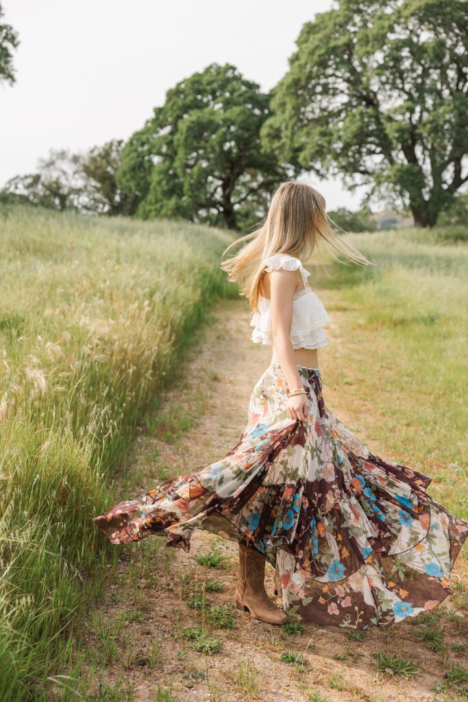 Senior girl posing on a dirt path between green rolling hills and oak trees in Danville, California wearing a boho floral maxi skirt and white ruffle top