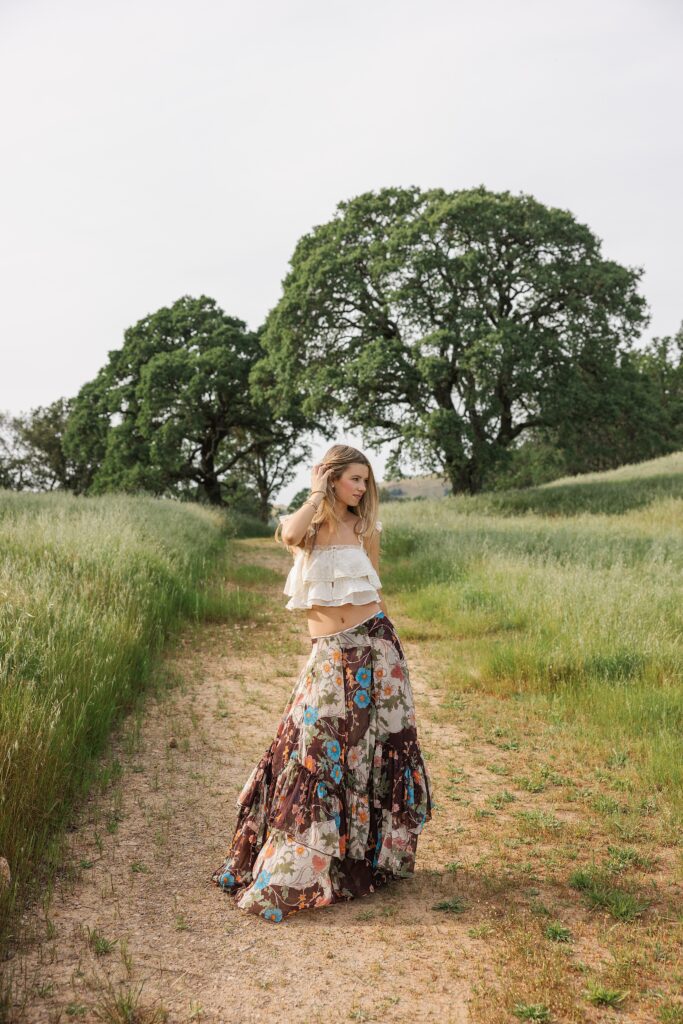 Senior portrait of a girl walking through a field in Danville, California wearing cowboy boots, a flowing floral skirt, and a white lace crop top with her hair blowing in the wind