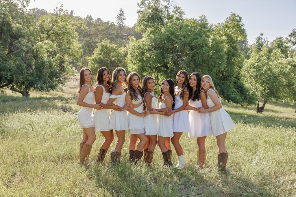 Group of senior girls wearing white dresses and cowboy boots, standing together in a sunlit grassy field in Danville, California for a senior BFF photo shoot 